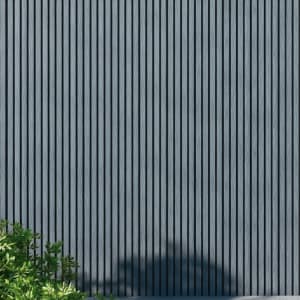 WPC slatted wall panels in gray tones, featuring uniform vertical slats and a natural wood grain texture, framed by green foliage and soft shadows. A minimalist architectural facade ideal for backgrounds, exteriors, design, and nature-inspired concepts.