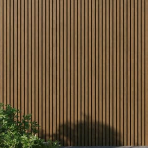 WPC slatted wall panels in oak tones, featuring uniform vertical slats and a natural wood grain texture, framed by green foliage and soft shadows. A minimalist architectural facade ideal for backgrounds, exteriors, design, and nature-inspired concepts.