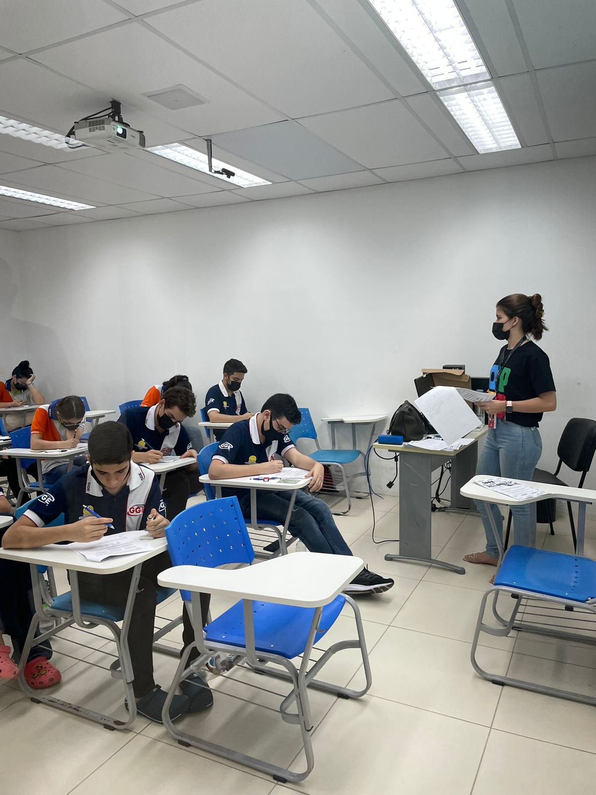 Sala com estudantes realizando a avaliação prática da OPEI em computadores. No canto direito, uma aluna aplica a prova, enquanto no meio e no canto esquerdo, alunos estão realizando a prova.