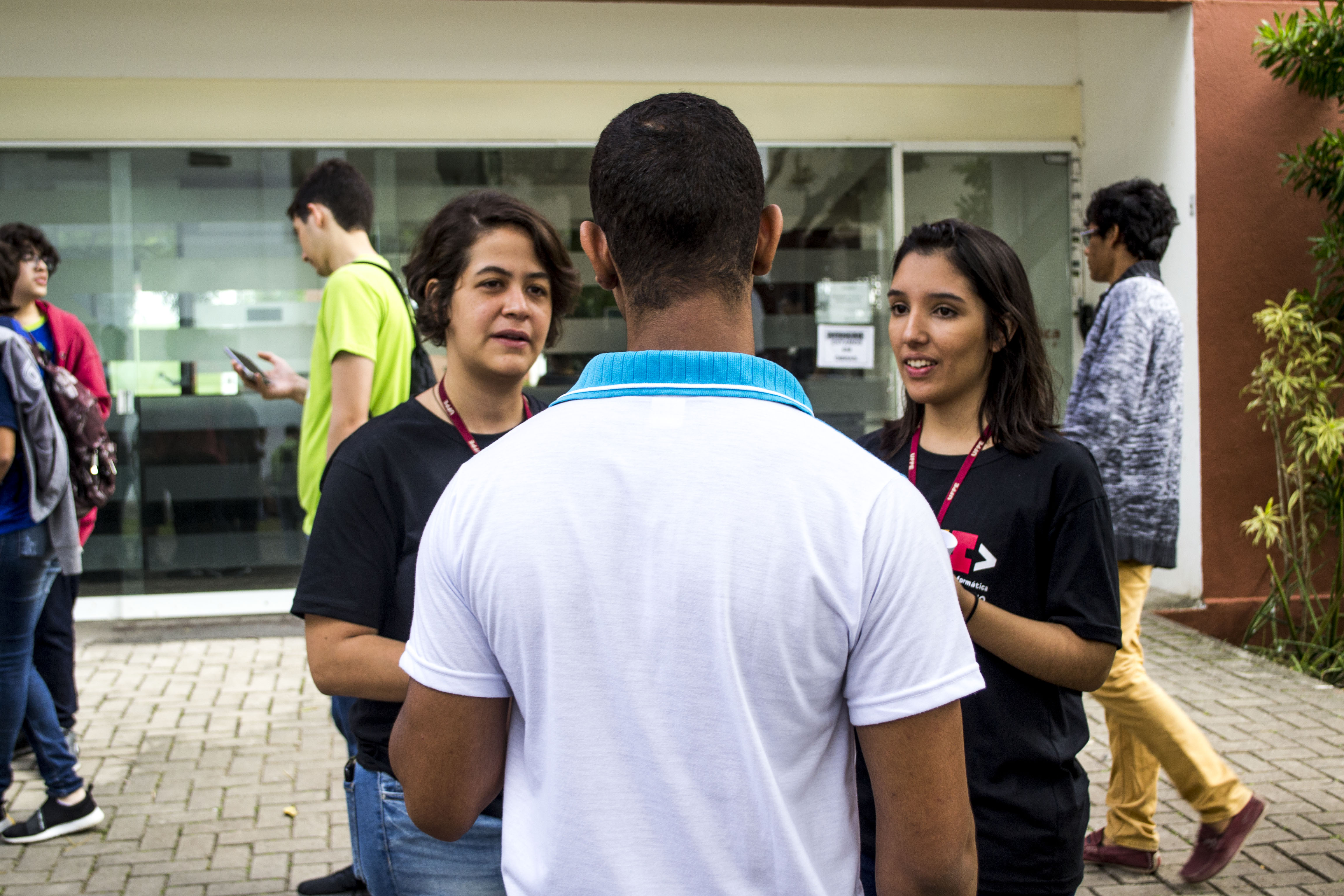 Duas petianas conversando com um participante da Olimpíada Pernambucana de Informática. Ao fundo, a entrada do Centro de Informática da Universidade Federal de Pernambuco e outras pessoas alunas.