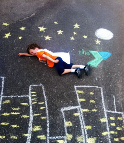 boy with cape laying on ground pretending to fly over a chalk mural of a city below a night sky with ymca brand - very cute