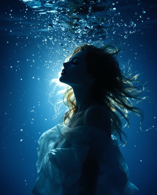 A woman underwater, lit from behind, bubbles rising around her