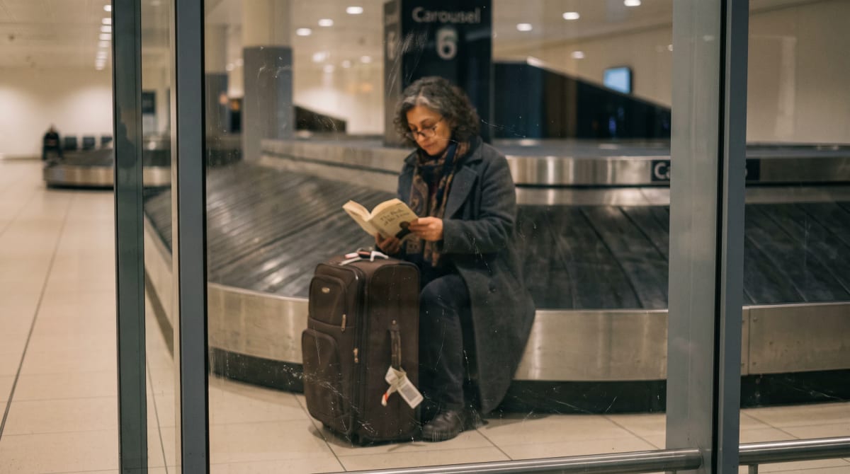 Traveler reading on a suitcase beside an empty airport baggage carousel