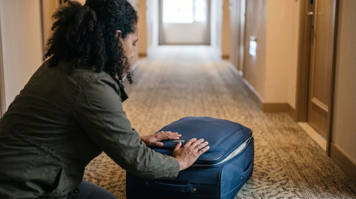 Traveler kneeling in a hotel hallway to zip a full suitcase closed