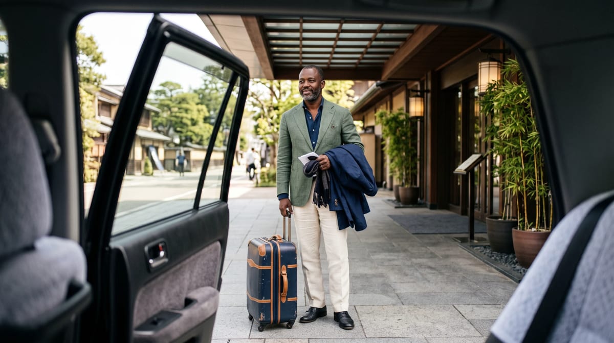 Traveler standing with a suitcase under a hotel canopy, seen through an open car door