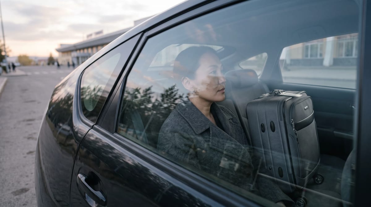 Traveler seated in a parked car at a quiet airport curb at sunrise, seen through the side window, suitcase beside
