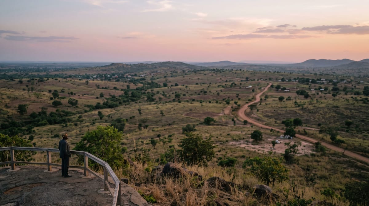 Man with hands in his pockets at a wooden rail on a roadside overlook, looking over a wide landscape at dusk