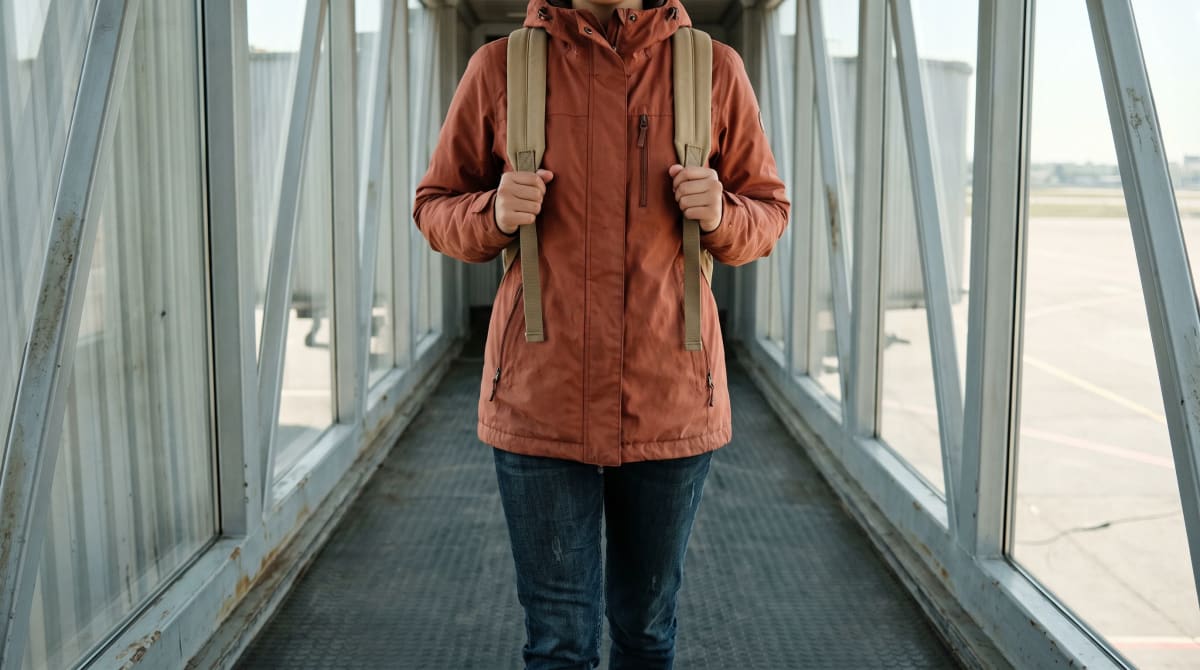Woman in a terracotta rain jacket holding her sand backpack straps as she walks toward the camera along an empty jet bridge