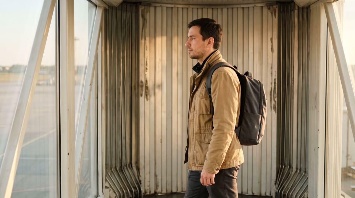 Man in a sand field jacket with a charcoal backpack walking past a side window along an empty jet bridge in golden light