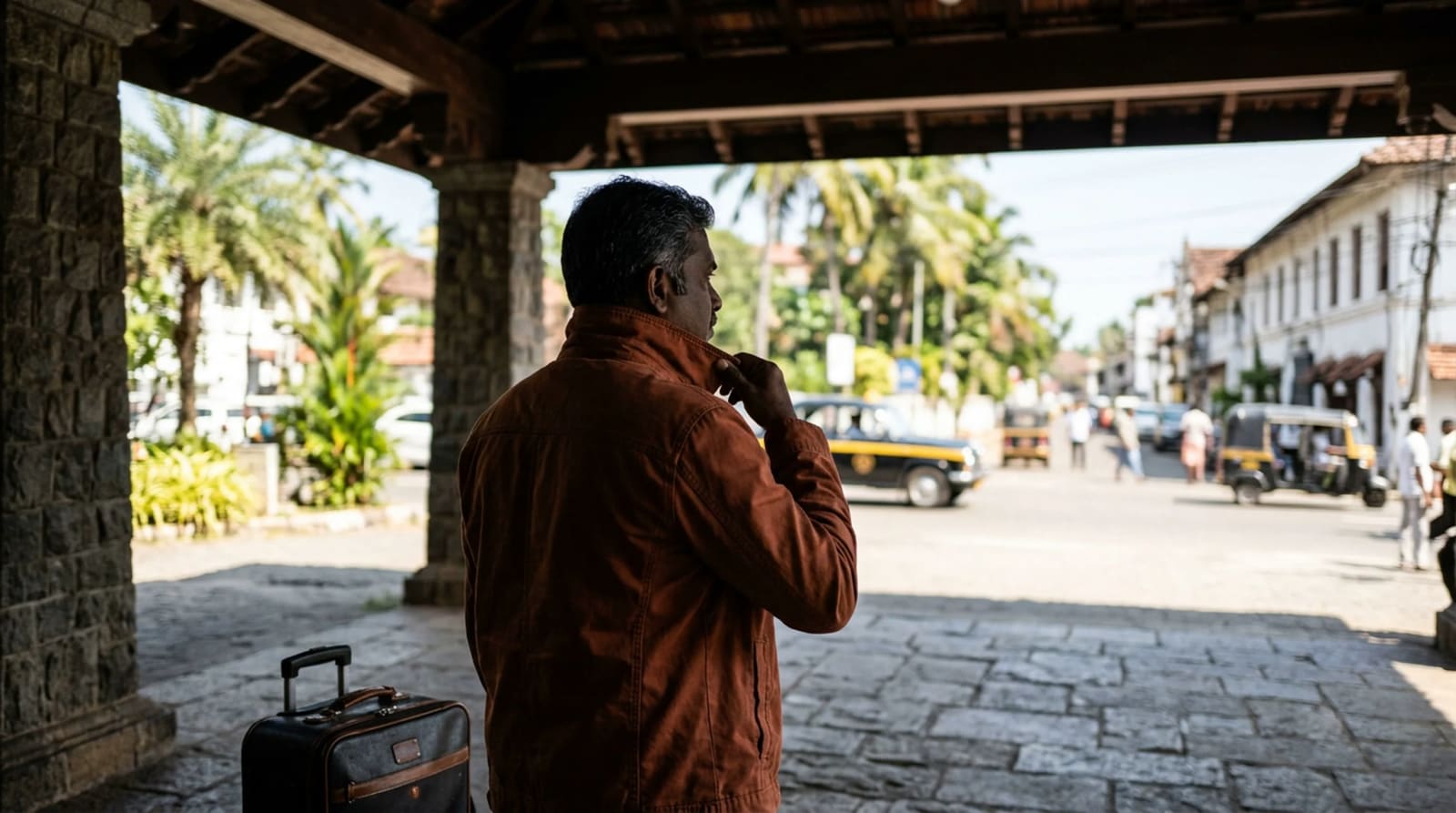 Traveler with one small suitcase standing just inside a hotel canopy looking out toward the bright drive