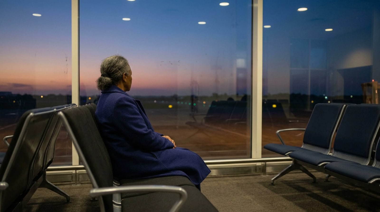 Woman in an indigo wool coat seated in a sparse airport gate row at dusk, facing a tall window over an empty apron