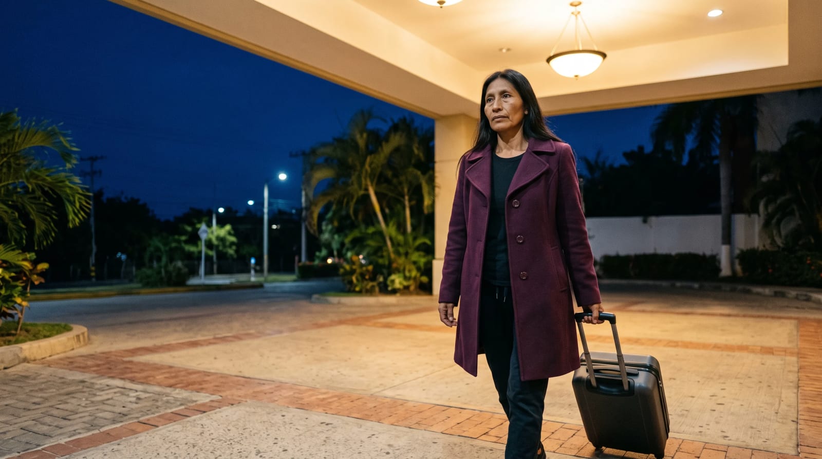 Woman in a plum wool coat pulling a small charcoal carry-on in under a lamplit hotel porte-cochère canopy at early night