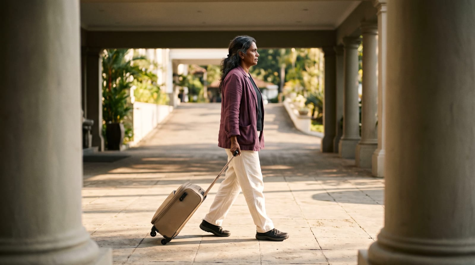 Woman in a plum linen jacket rolling a sand carry-on in from the drive under a hotel porte-cochère canopy in afternoon light