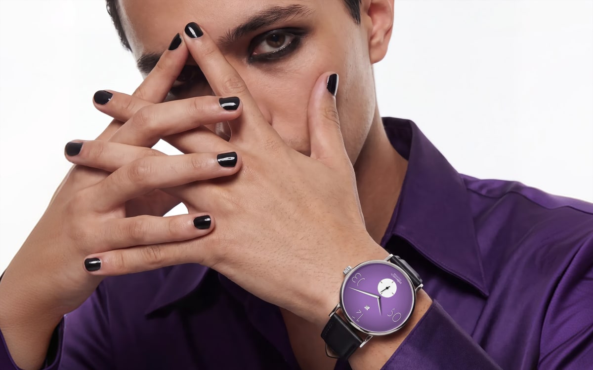Manos entrelazadas con las uñas pintadas de negro y el Marcha 383 Malasaña, esfera lavanda, en la muñeca de una camisa morada