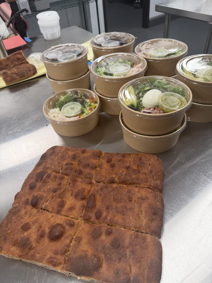 Stacked lidded pickup bowls of salad next to a tray of dimpled focaccia