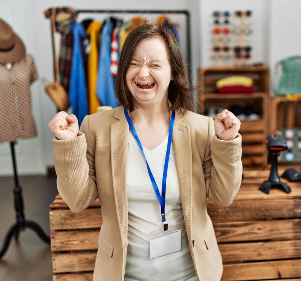 Young down syndrome woman working as manager at retail boutique excited for success with arms raised and eyes closed celebrating victory smiling. winner concept.