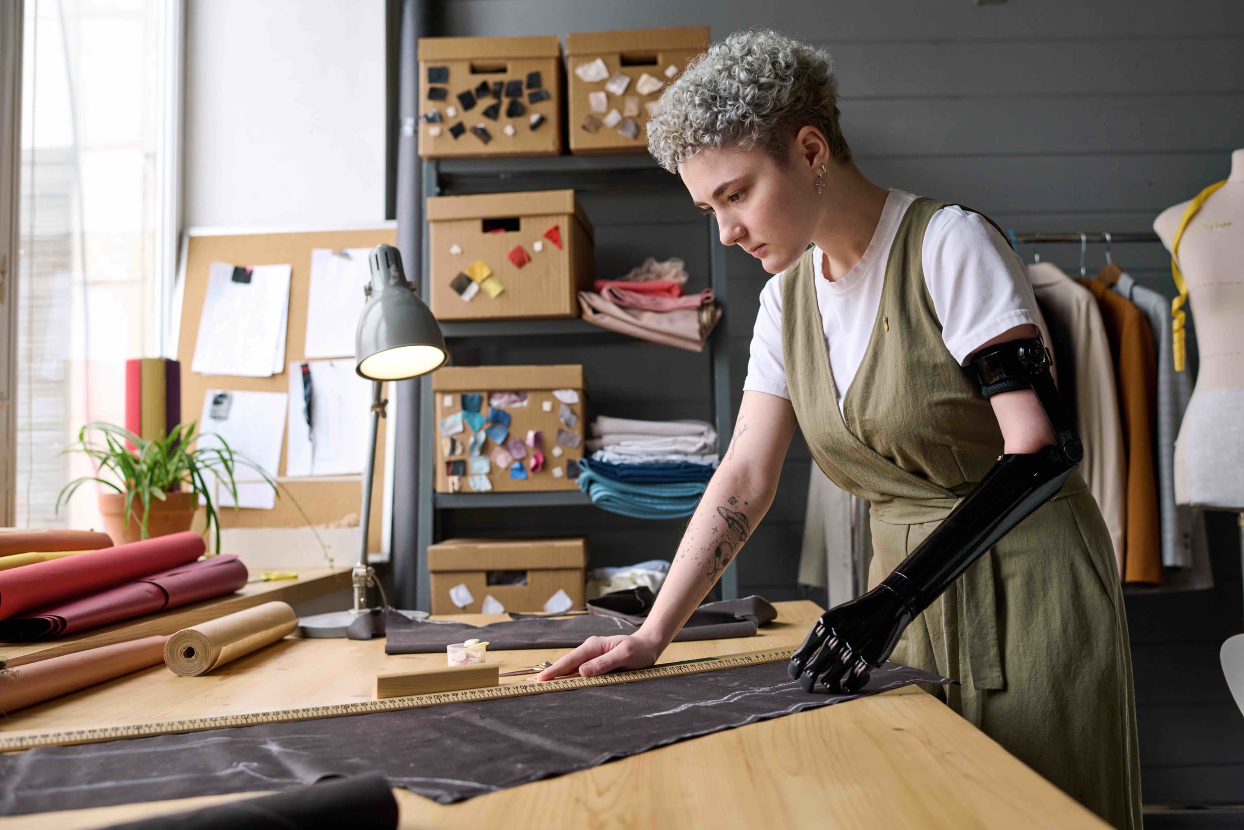 Training services at MovingMood. Young female tailor with black arm prosthesis standing by table in workshop or studio creating new clothes