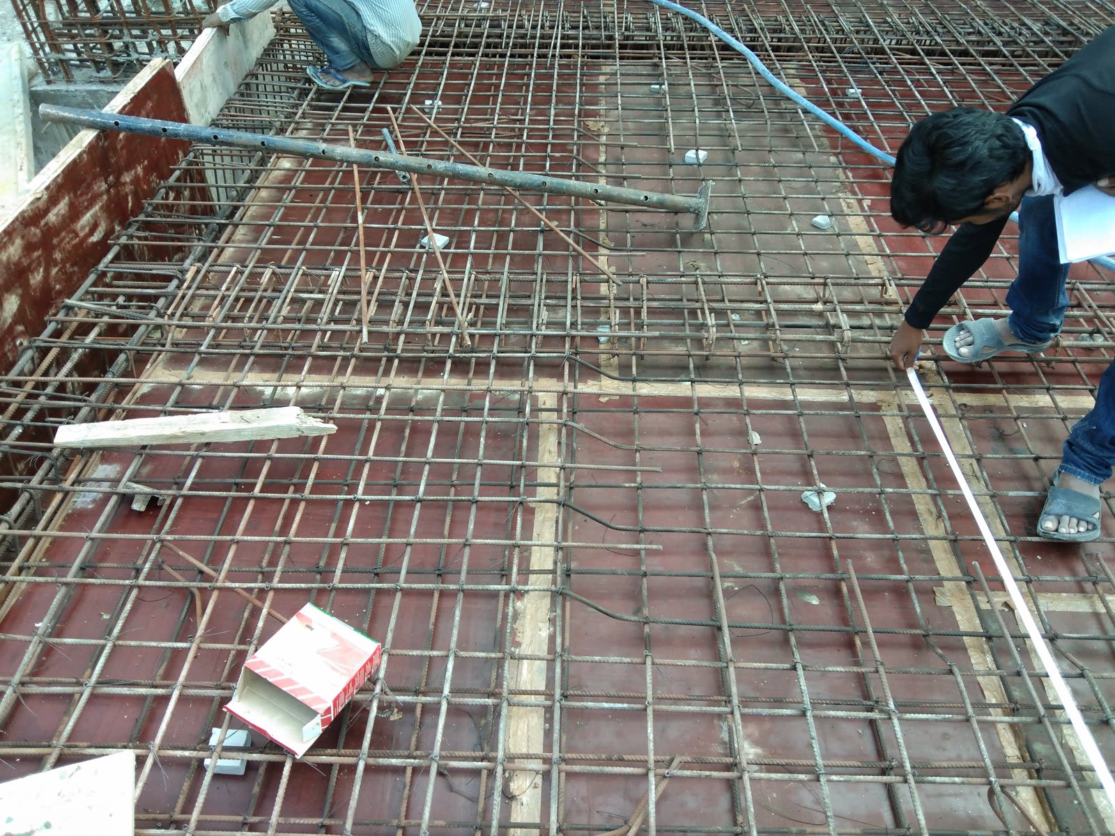 Steel reinforcement mesh laid over red formwork, with a person measuring bar spacing with a tape