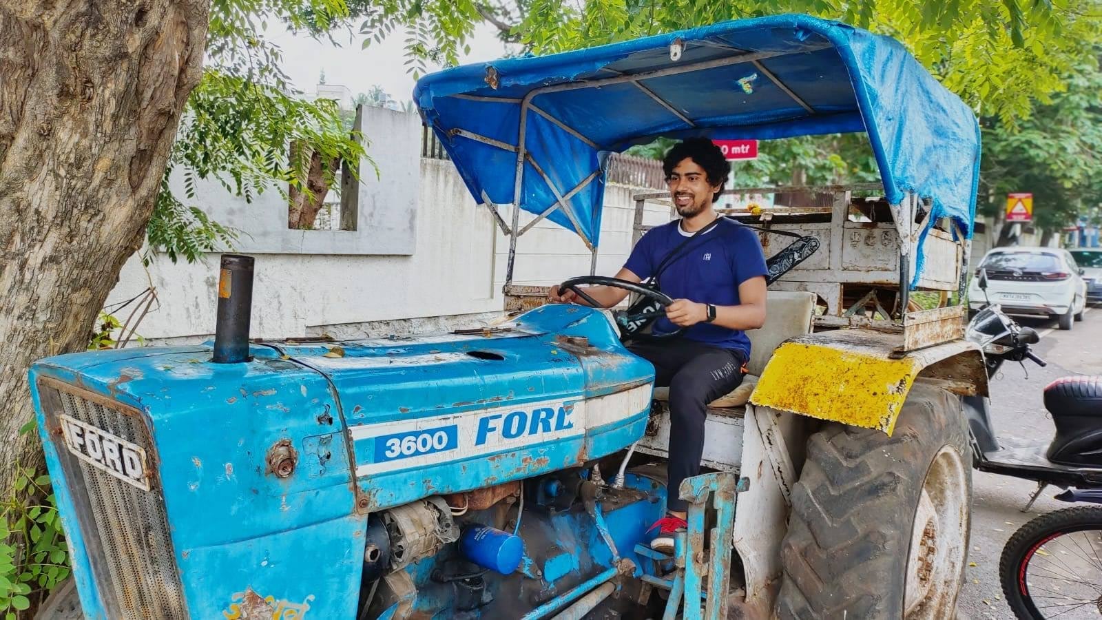 Sagar smiling at the wheel of an old blue Ford tractor parked under a tree