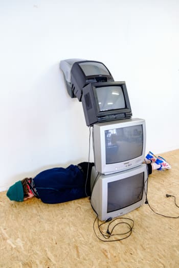 Kunstenaar licht plat op zijn buik onder kunstwerken die bestaan uit 5 tv's die schuin op elkaar zijn geplaatst en vervaarlijk tegen de muur aanleunen waardoor ze een halve boog vormen.