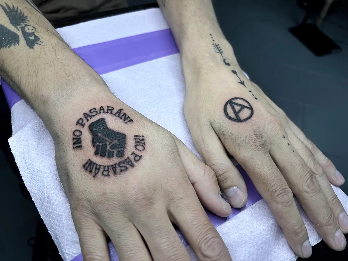 Tatuaż na dłoniach z zaciśniętą pięścią i napisem No Pasaran oraz symbolem anarchii