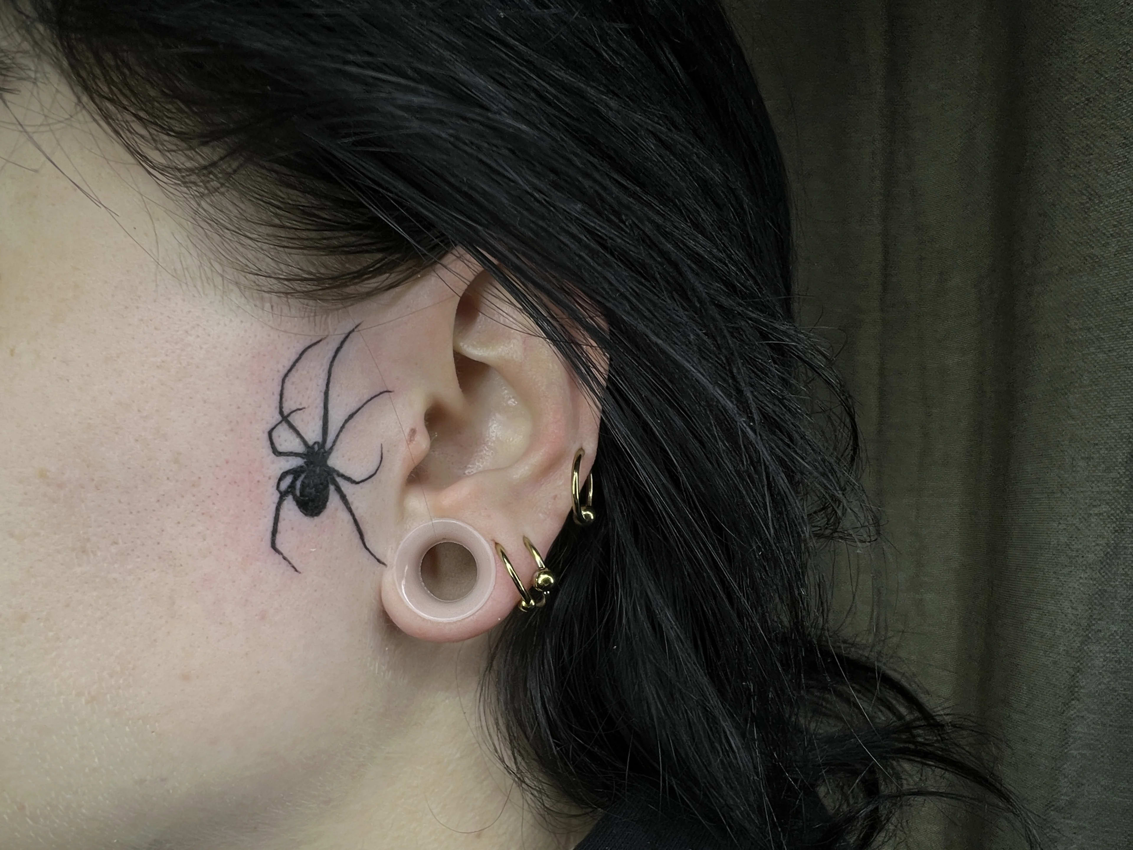 Pequeño tatuaje de una araña en la piel, justo al lado de la oreja, en estilo minimalista.