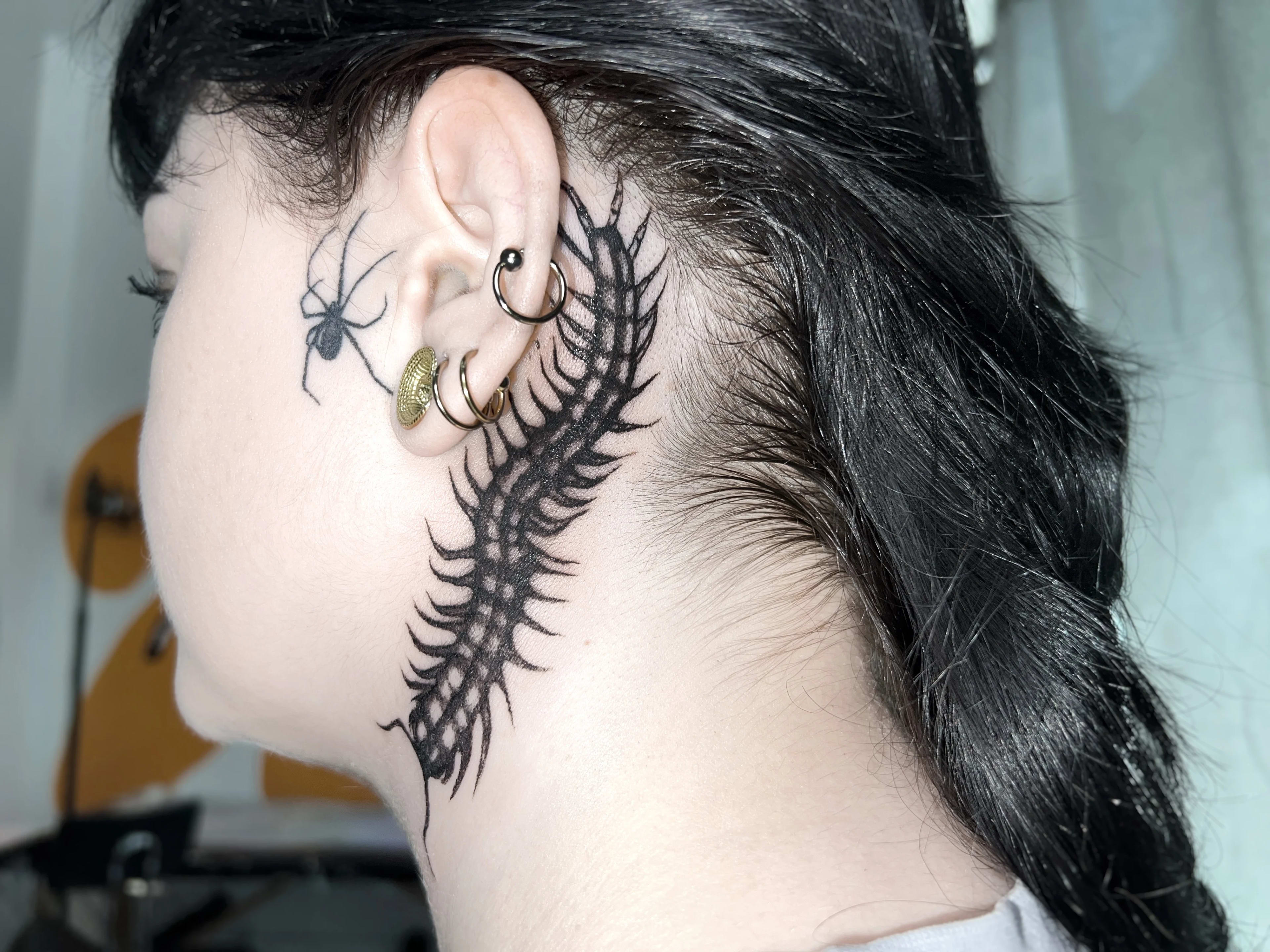 Tatuaje de una escolopendra detrás de la oreja y una araña en la mejilla, en estilo blackwork.