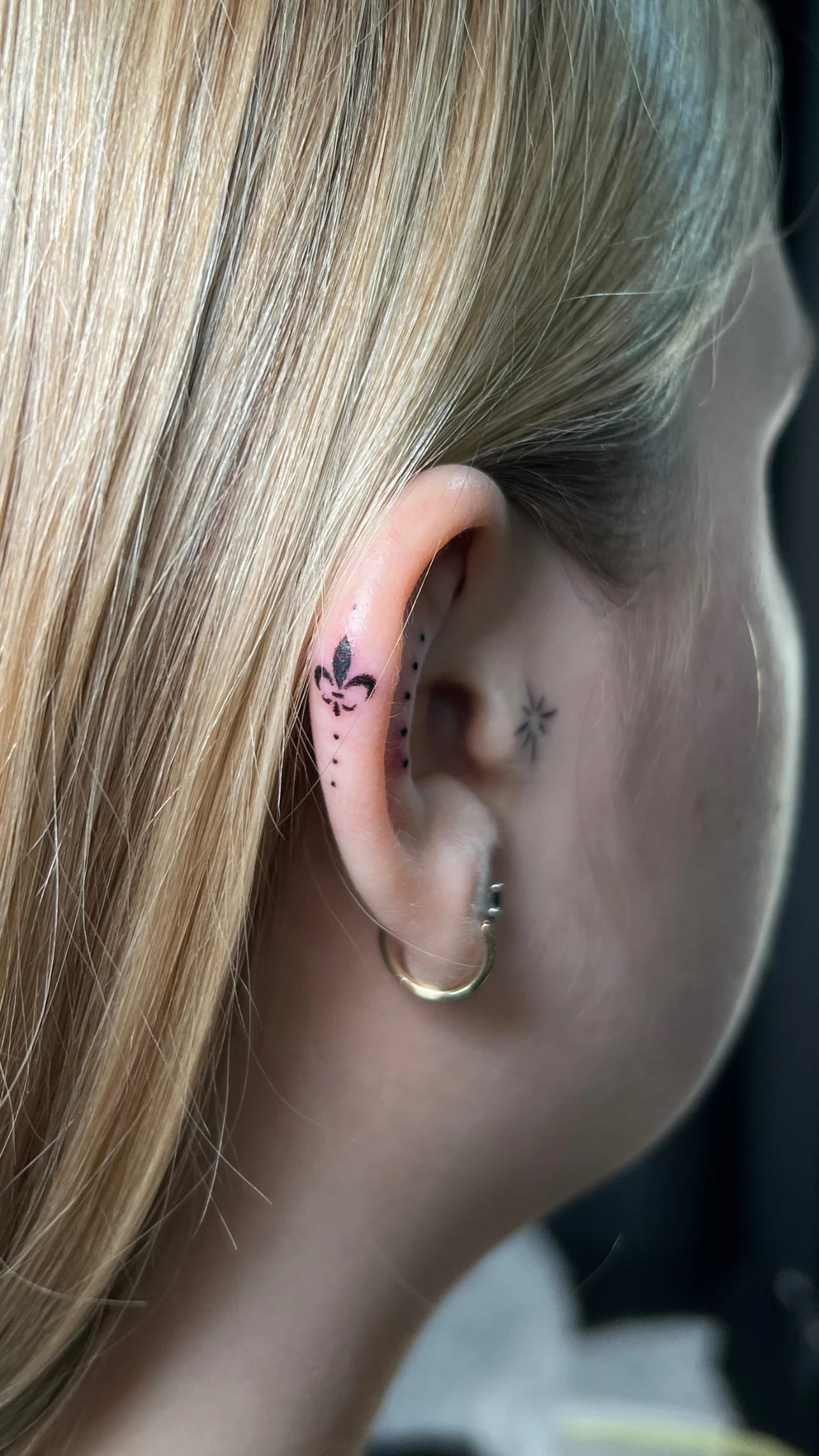Pequeño tatuaje con una flor de lis y puntos en el cartílago de la oreja.