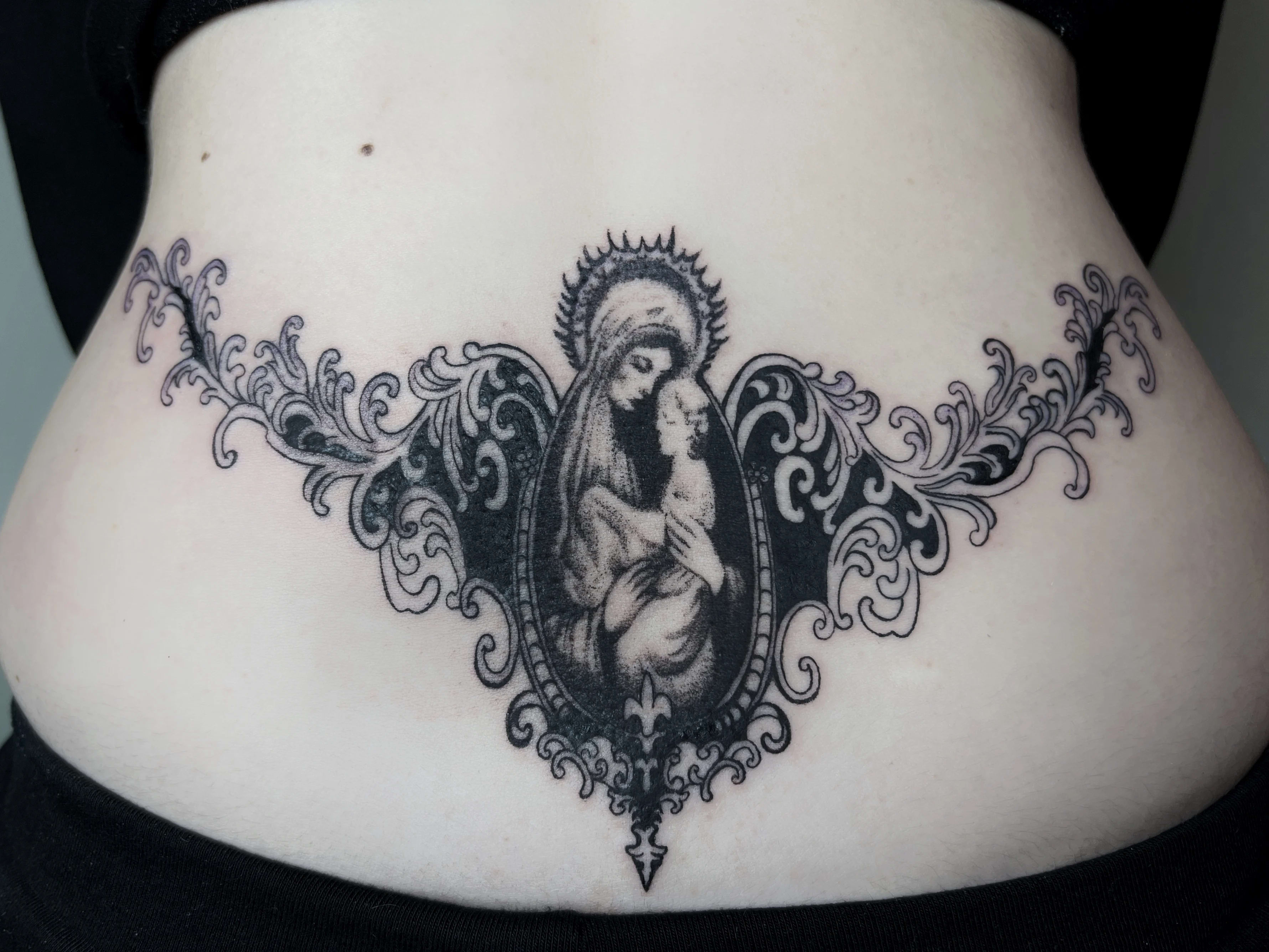 Tatuaż z Madonną z Dzieciątkiem w ozdobnej ramie na dolnej części pleców w stylu blackwork.