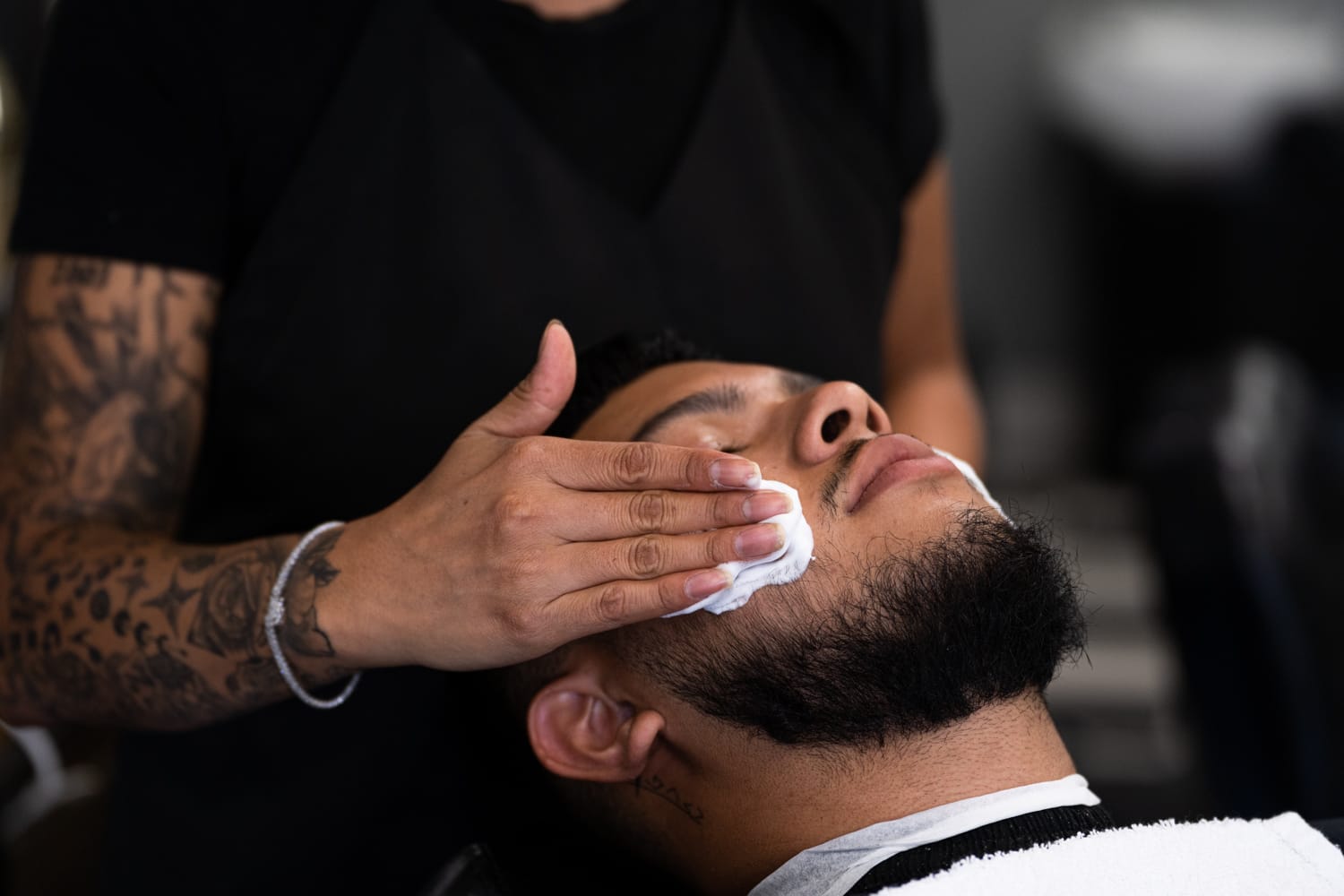 A man leans his head back as the barber behind him gently applies shaving cream to his cheeks.