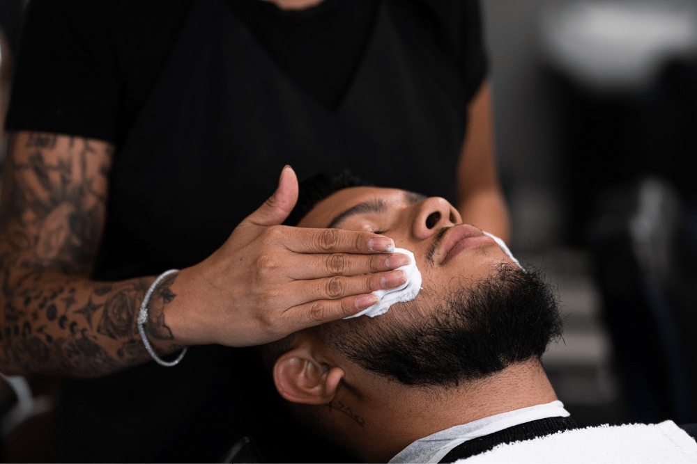 A man leans his head back as the barber gently applies shaving cream to his face.