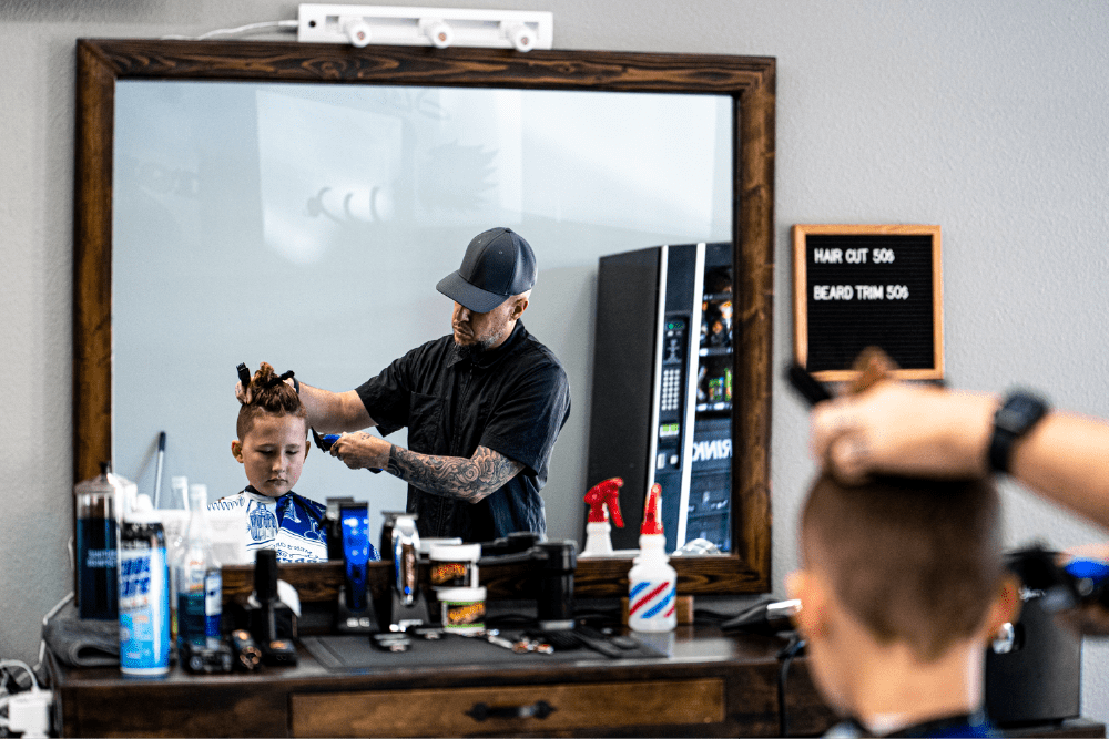 Viewed from a reflection in a mirror, a boy sits in a chair while the barber gives a haircut.