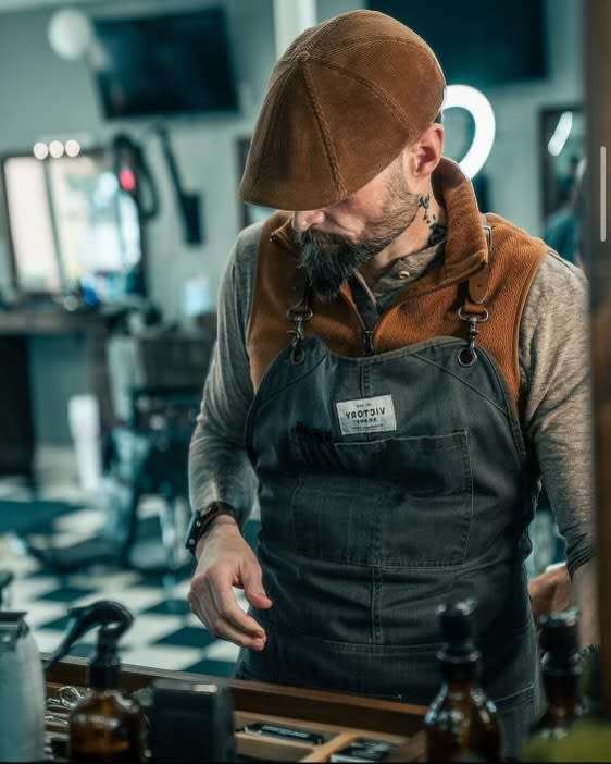 A man with a serious expression and hat sits in a barber chair with his hands folded.