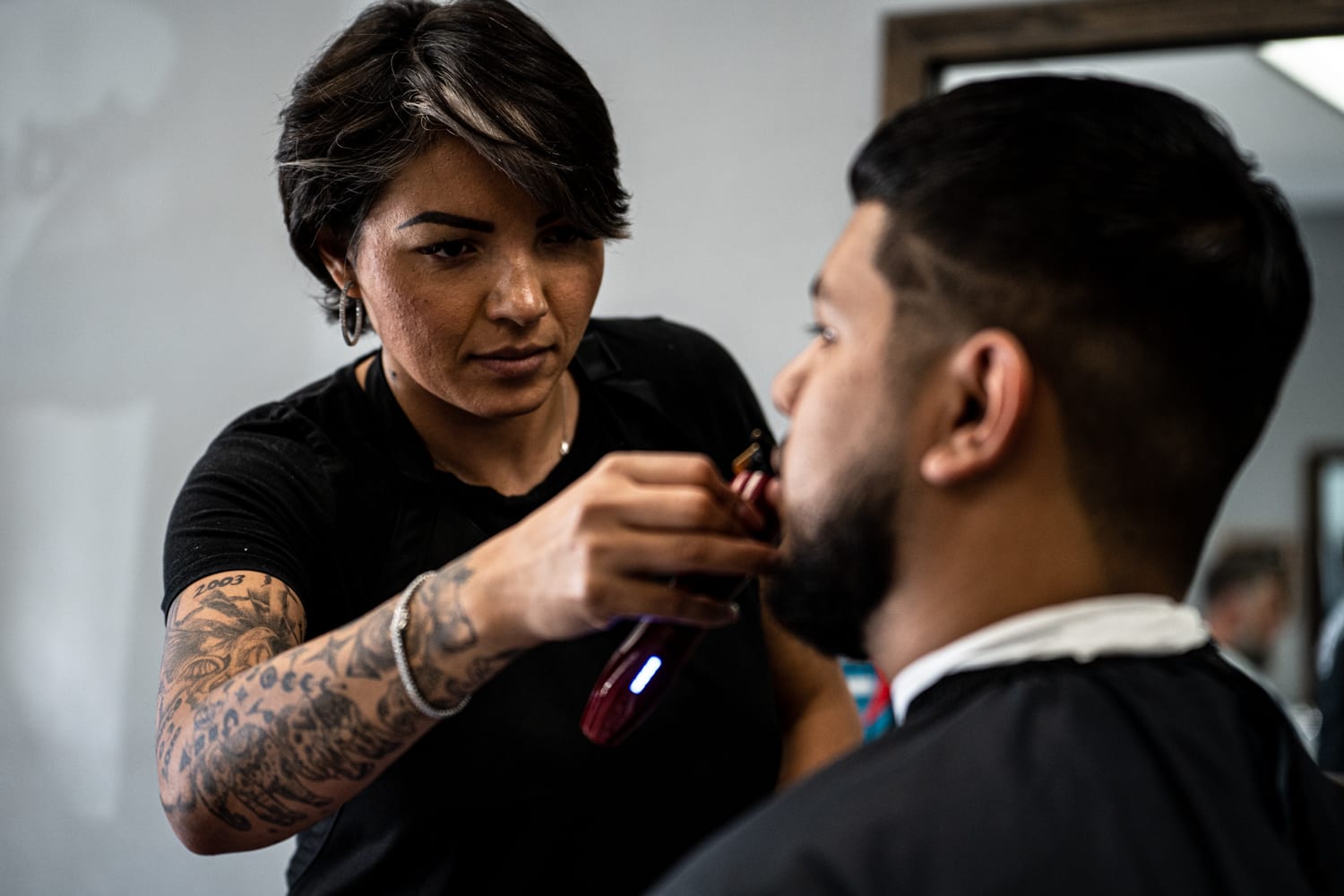 A barber uses her electric razor to trim the sides of a man's moustache.