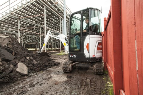 A Bobcat E20 Compact Excavator Maneuvers Between a Fence and Aluminum Bleachers While Digging Dirt 