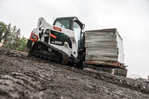 A Bobcat T595 Compact Track Loader Carries a Heavy Pallet of Block Across a Flat Dirt Surface