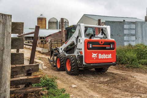 A Bobcat S595 skid-steer loader with root grapple attachment drives down lane heading toward farm buildings