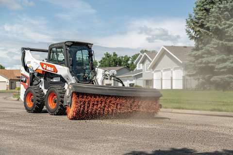 Bobcat skid-steer loader using an angle broom attachment to sweep debris from a paved street.