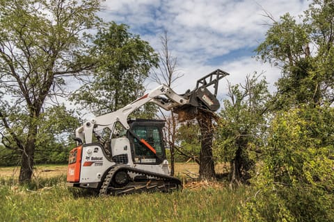 Landscaper Operating T870 Compact Track Loader With Forestry Cutter Attachment To Cut Through Tree