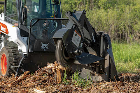 Bobcat loader with high-flow hydraulics powering a stump grinder attachment in a wooded area.
