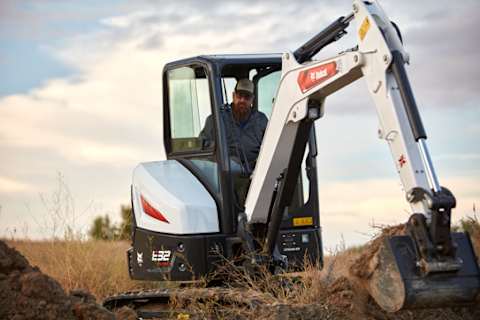 Operator in a Bobcat E32 Compact (mini) Excavator Digs Hole in Filed With Clouds in Background.
