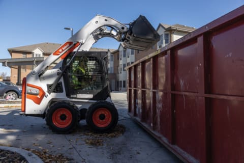 Operator Using Bobcat Skid-Steer Loader With Bucket Attachment To Dump Debris Into Dumpster