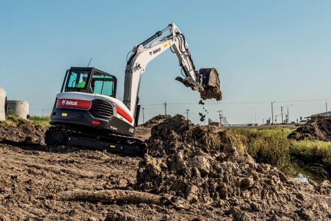 A Bobcat E85 compact excavator lifts a full bucket of dirt at a jobsite.