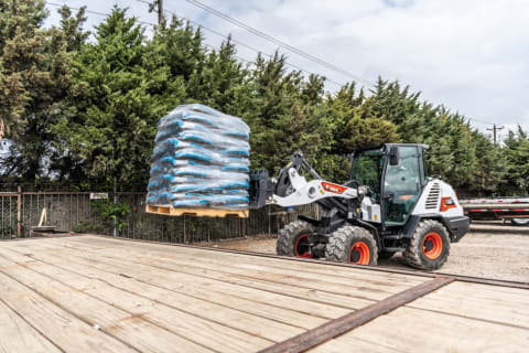Bobcat L85 Compact Wheel Loader With Pallet Fork Attachment Moves Material On To A Trailer.
