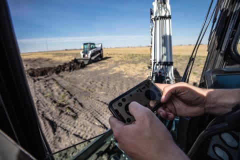 A Worker Uses Bobcat MaxControl to Operate a Platinum Bobcat Compact Track Loader From the Cab of a Nearby Excavator