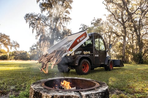 A Toolcat UW56 Dumps Firewood in a Green, Wooded Area Near a Firepit