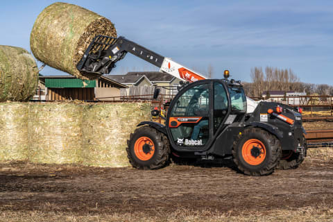 A Bobcat TL619 lifting a hay bale off of a hay bale pile using a bale fork