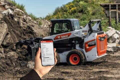 An individual’s hand, holding a cellular phone with a Bobcat app open on the screen, appears in the foreground of a photo while a Bobcat skid-steer loader works in the background. 
