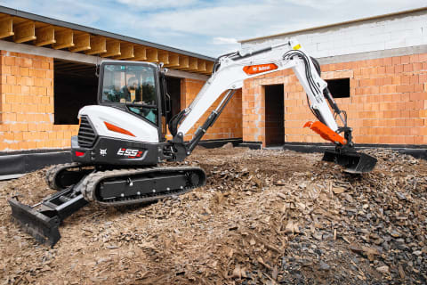 Een mannelijke bestuurder verplaatst met een Bobcat-minigraafmachine voorzien van een lange arm puin op een bouwlocatie. 