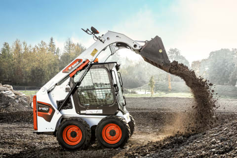 Una cargadora compacta Bobcat con los brazos extendidos vertiendo rocas pequeñas sobre una montón mientras realiza trabajos de jardinería en un día soleado.